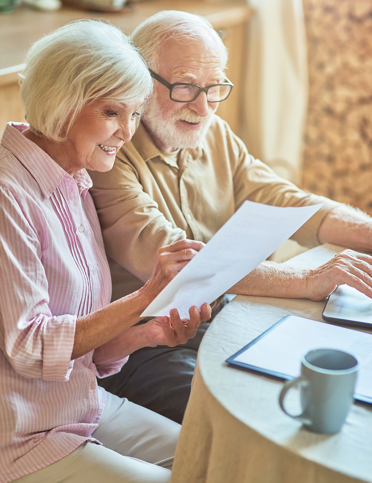 Elderly couple reviewing documents at a table; woman points to paper, man uses laptop, mug on table.